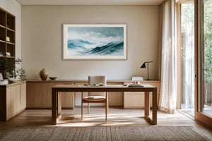 Framed watercolor mountain print above a wooden desk beside tall windows.