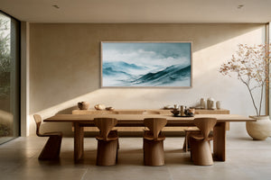 Large framed blue mountain watercolor above a long wooden dining table and sculptural chairs.