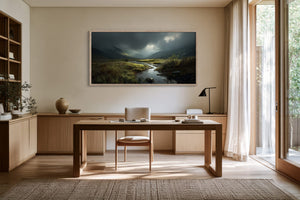 Framed mountain valley stream artwork above a wooden desk beside tall glass doors.