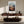 Light wood-framed mountain valley artwork above a dark wood sideboard in a sunlit seating area.