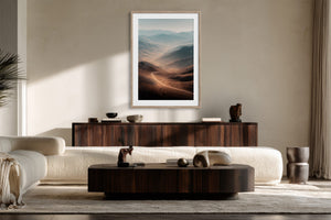 Light wood-framed mountain valley artwork above a dark wood sideboard in a sunlit seating area.