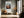 Light wood-framed mountain valley artwork above a cushioned bench beside tall windows.