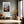 Light wood-framed mountain valley artwork above a cushioned bench beside tall windows.