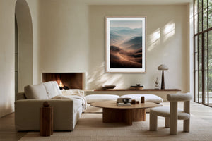 Light wood-framed mountain valley artwork above a fireplace and cream seating near grid windows.