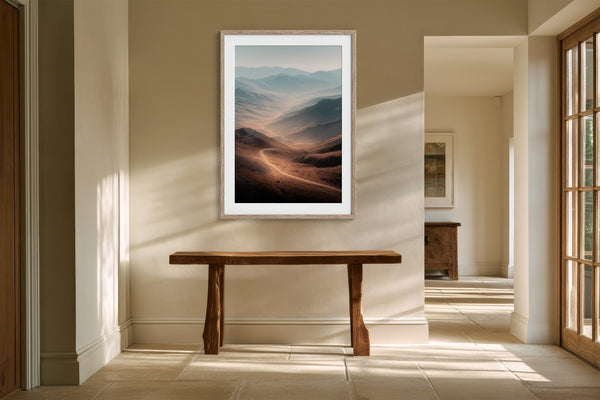 Light wood-framed mountain valley artwork above a rustic console table in a sunlit hallway.